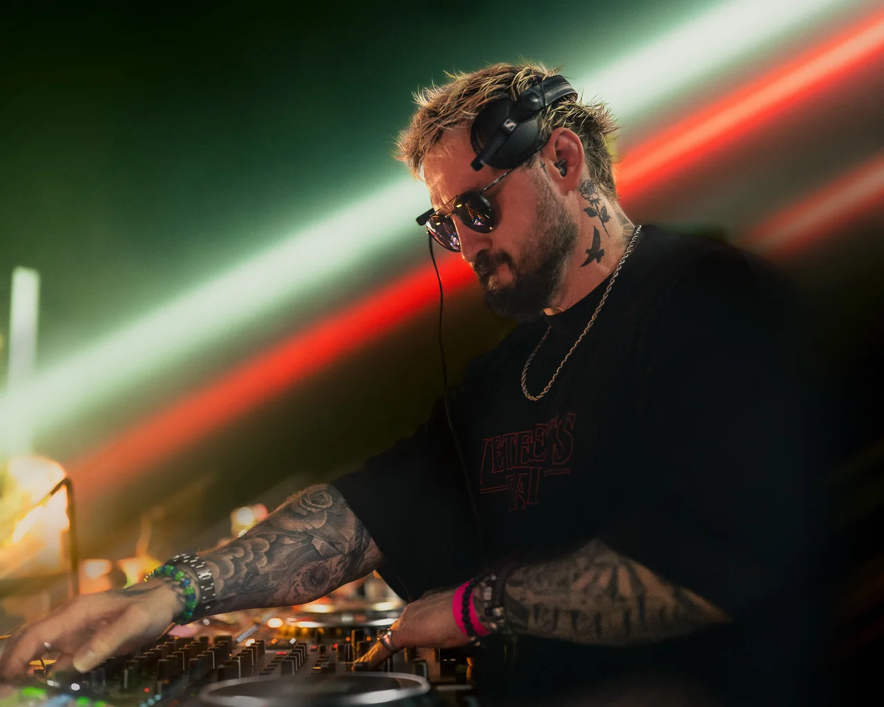DJ in sunglasses performing beneath diagonal red and green beams.