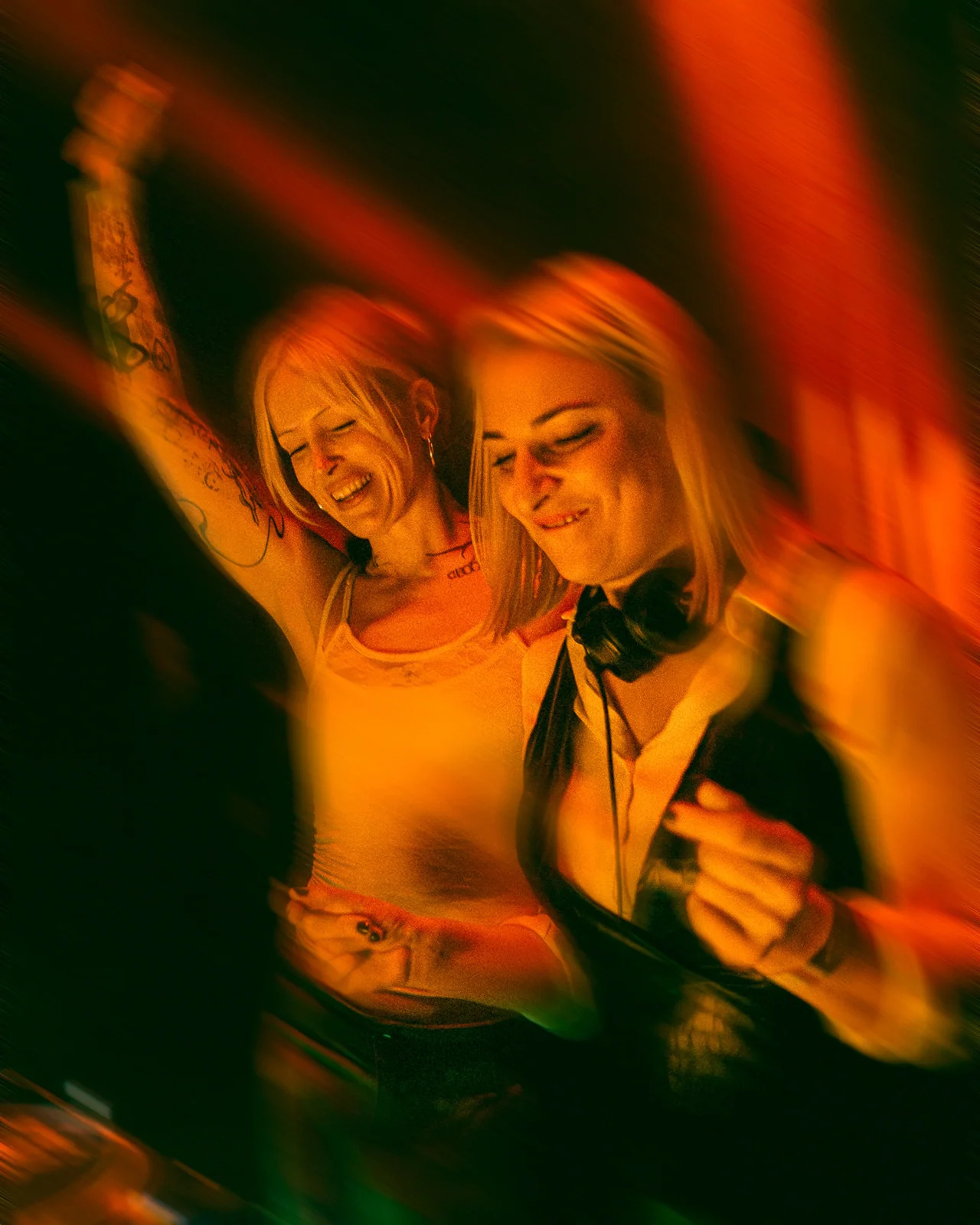 Two women sharing a quiet moment under warm orange club light.