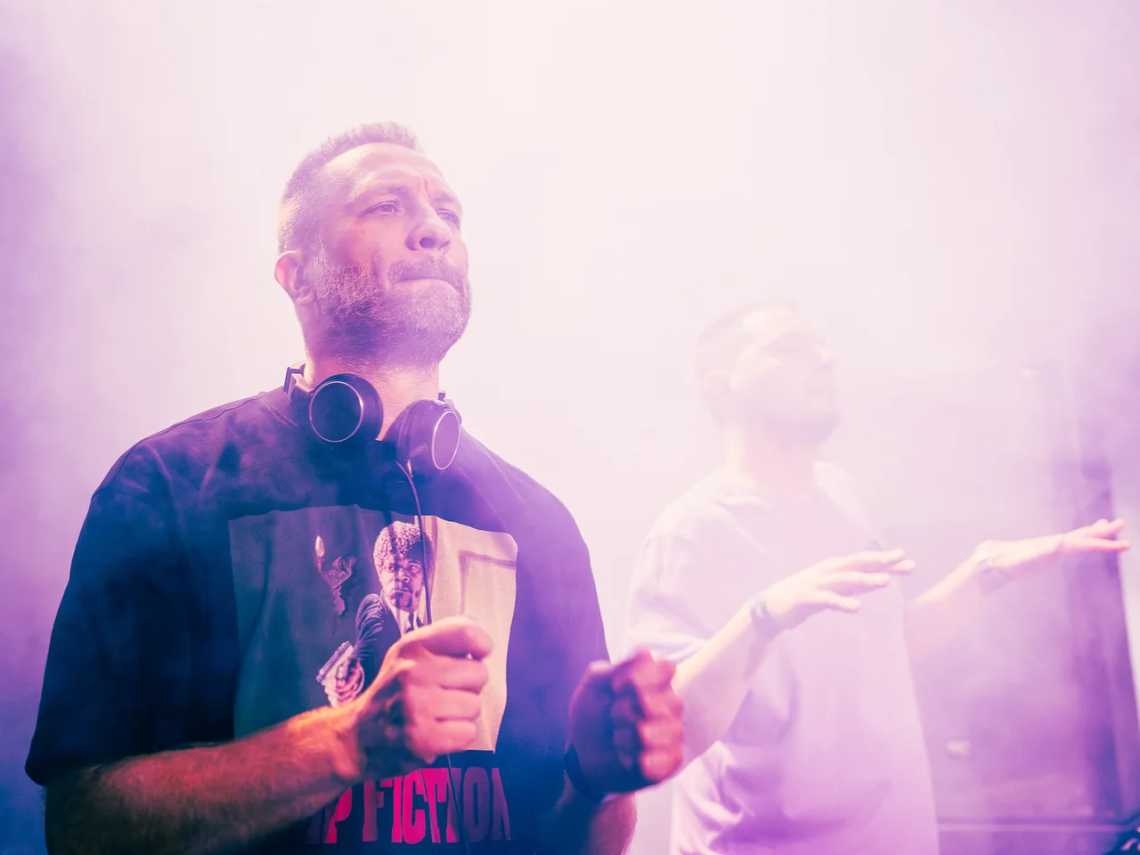 DJ holding a drink in the booth beneath soft pink haze.