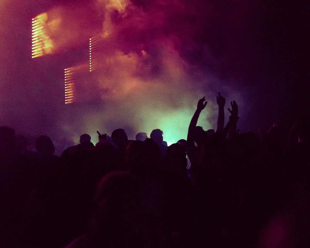 Audience silhouettes and raised hands beneath magenta stage light.