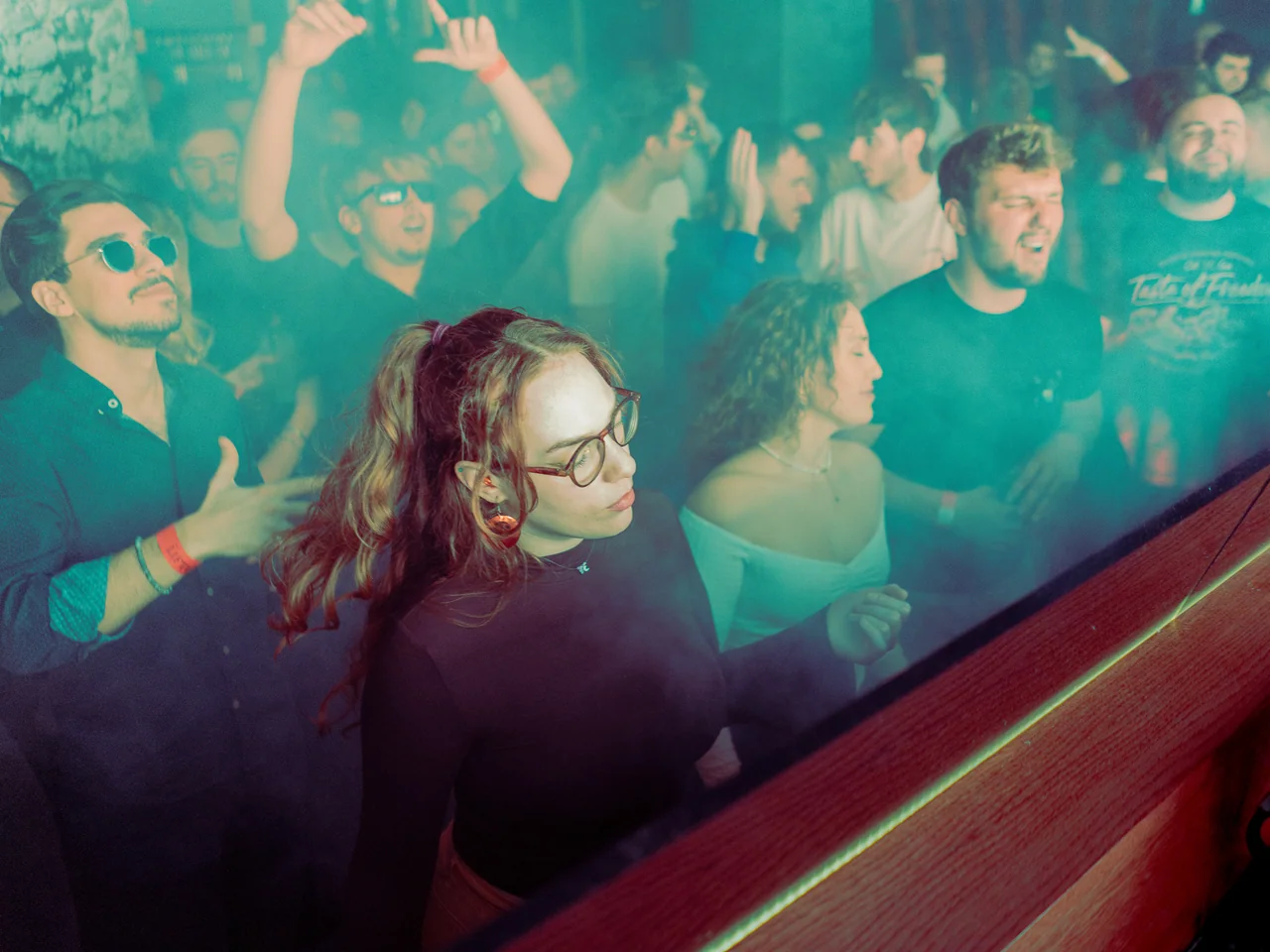 DJ with glasses framed by the crowd and turquoise haze.