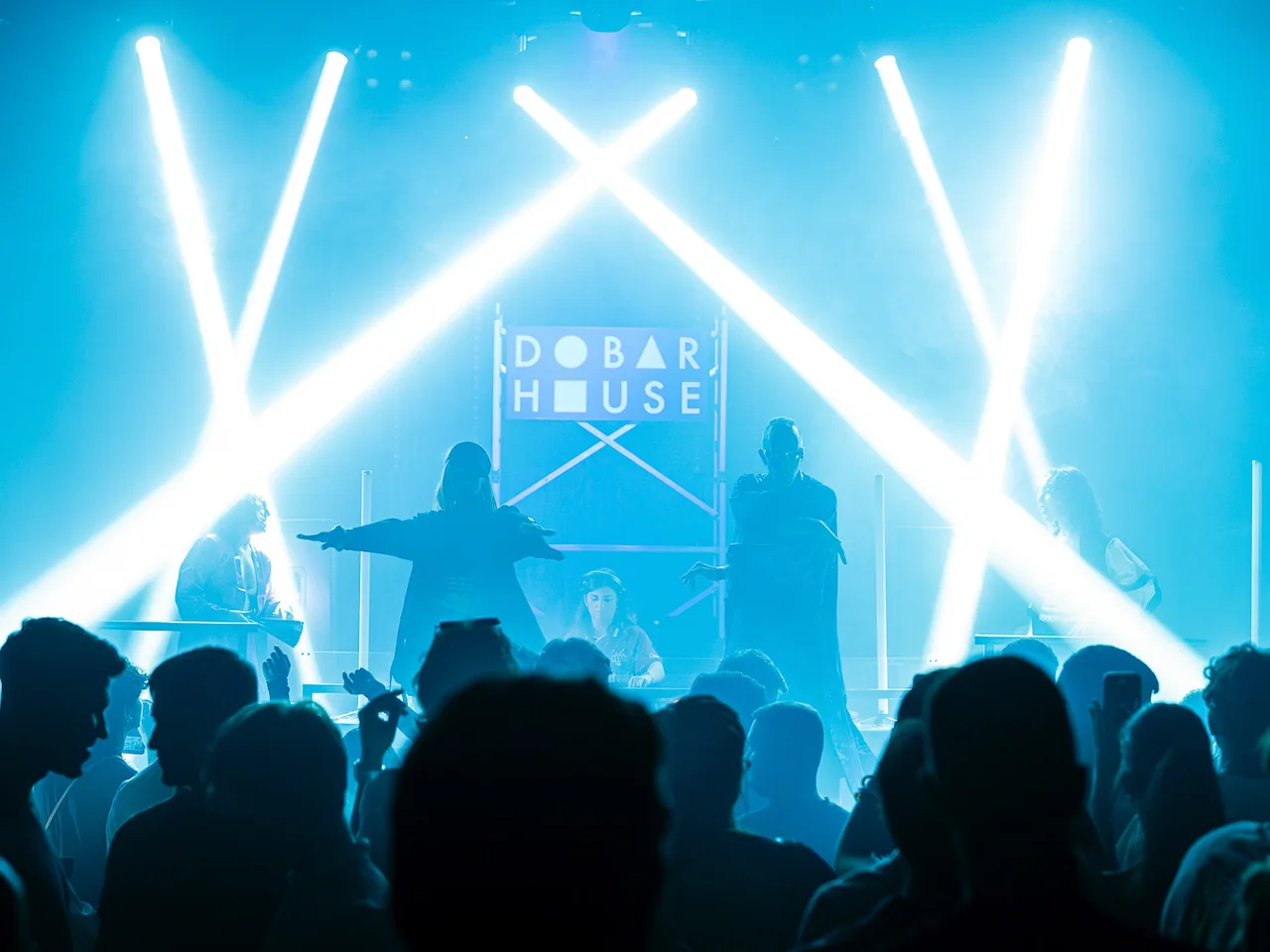 House-music stage silhouetted by bright white and cyan beams.
